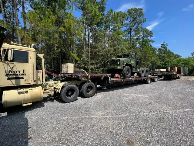 Tan tractor truck pulling a lowboy trailer loaded with a green truck